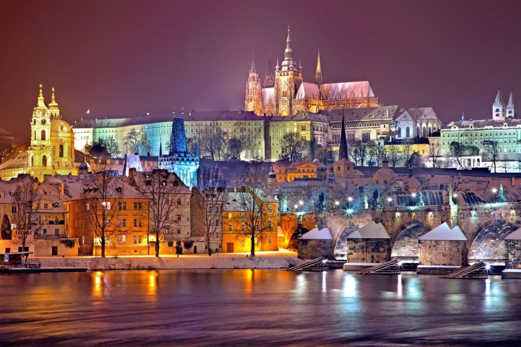 Castello di Praga illuminato di notte, con il fiume e i tetti innevati in primo piano